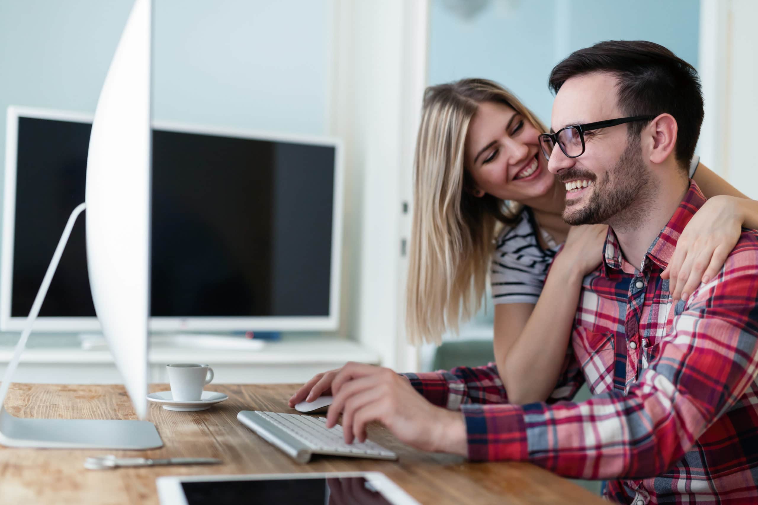 A younger man sitting on his computer with his iPad on the desk to his left. He is typing on his computer and his girlfriend has her arms around him. They are both smiling