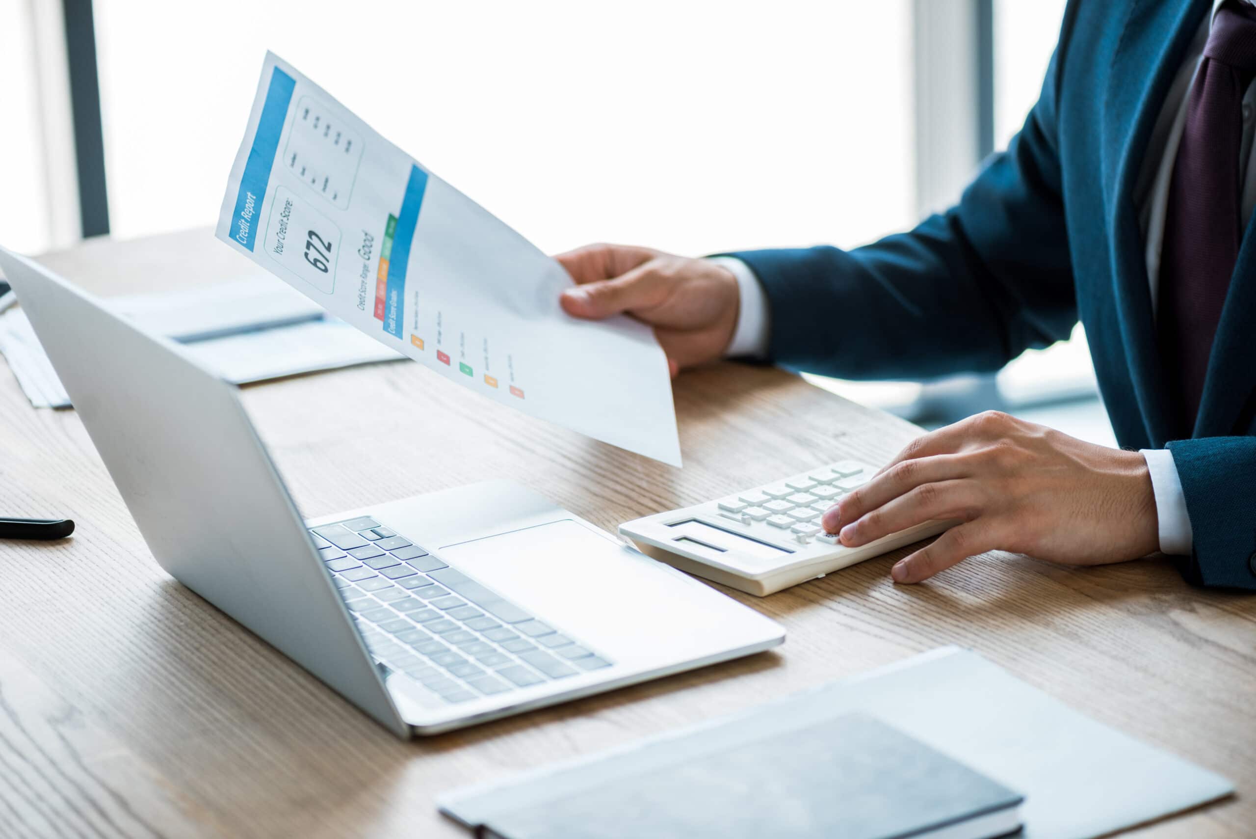 A cropped photo of a man holding his credit report doing calculations on his calculator. He has his laptop in front of him and is wearing a blue suit.